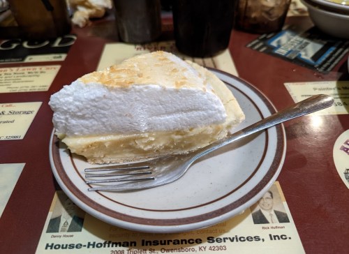 A slice of lemon meringue pie on a plate with a fork, resting on a table covered with menus and advertisements.