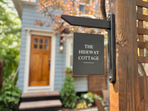 A sign reading "The Hideaway Cottage" hangs on a wooden post, with a cozy cottage in the background.