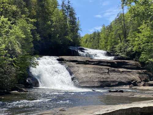 A serene waterfall cascades over rocky ledges, surrounded by lush green trees under a clear blue sky.