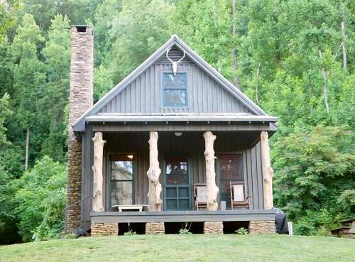 A rustic wooden cabin with a stone chimney, surrounded by lush greenery and featuring unique log pillars on the porch.