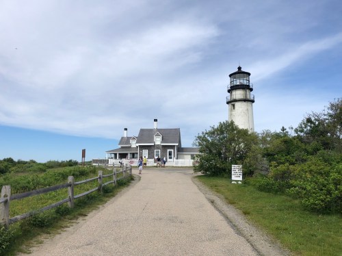 A lighthouse beside a white building, surrounded by greenery and a gravel path under a blue sky.