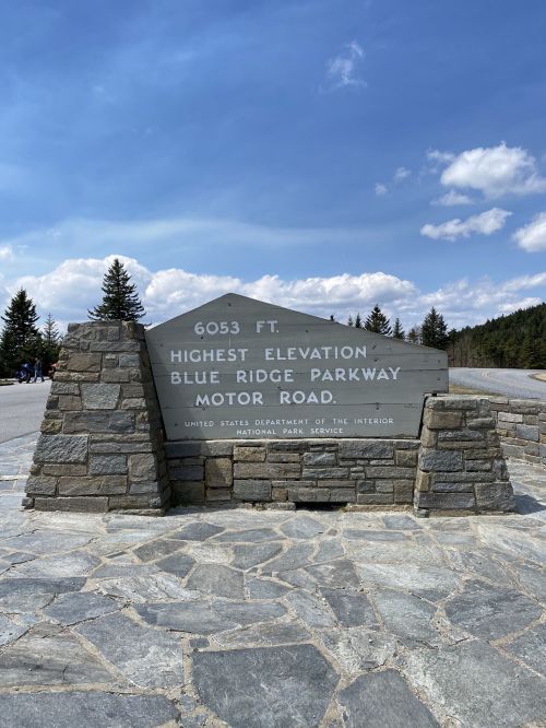 Sign marking the highest elevation on the Blue Ridge Parkway, 6053 ft, with trees and a blue sky in the background.