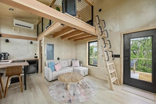 Cozy interior of a modern tiny house featuring a loft, sofa, wooden ladder, and large windows with a view.