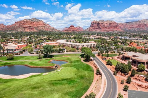 Aerial view of a golf course with a pond, surrounded by lush greenery and red rock formations under a blue sky.