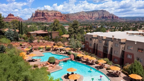 A scenic view of a resort pool surrounded by lush trees and mountains under a partly cloudy sky.