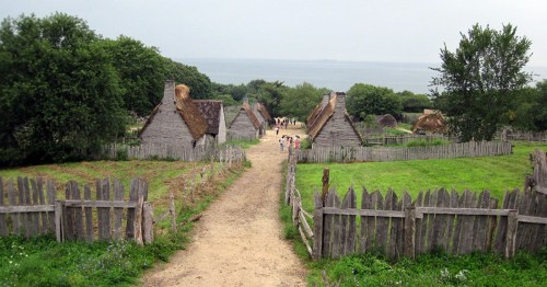 A scenic view of a historic village with thatched-roof houses, surrounded by greenery and a distant body of water.