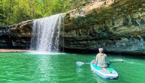 A person in a kayak paddles near a waterfall in a lush, green landscape with clear turquoise water.