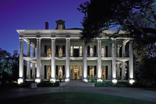 A grand, illuminated mansion with tall columns, set against a twilight sky, surrounded by lush greenery.