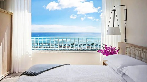 Bright hotel room with a bed, large window showcasing a blue ocean view, and a touch of pink flowers.
