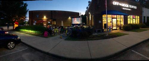 Outdoor dining scene at Off the Hook Seafood restaurant, with a large screen showing a movie and patrons enjoying the evening.
