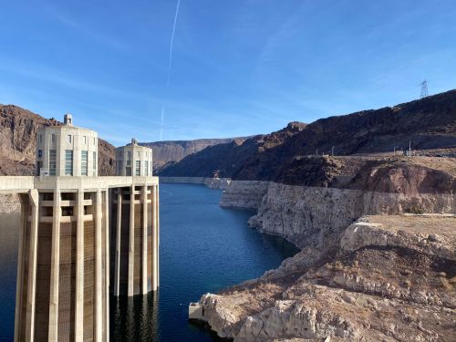 View of a dam with two towers, surrounded by rocky cliffs and a calm blue lake under a clear sky.