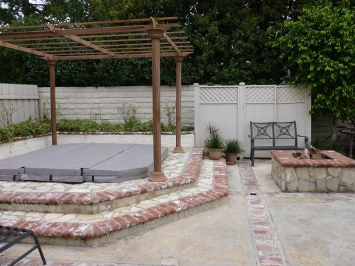 A backyard patio with a pergola, brick pathways, a bench, and a hot tub cover surrounded by greenery.