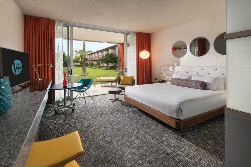 Modern hotel room with a queen bed, desk, colorful chairs, and large windows opening to an outdoor view.