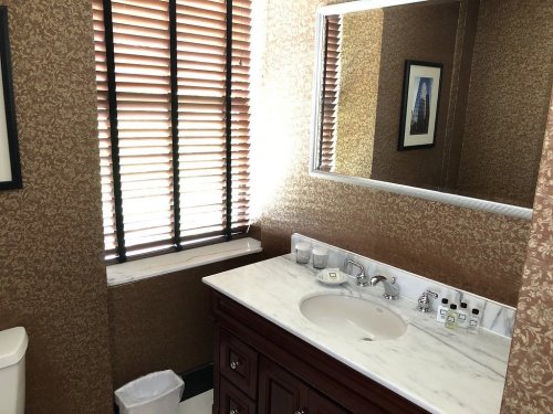 A bathroom with a marble countertop, wooden vanity, mirror, and window with blinds, featuring decorative wallpaper.