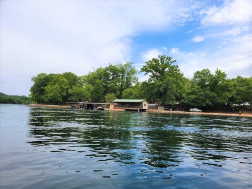A serene lake scene with a house and dock surrounded by lush green trees under a partly cloudy sky.