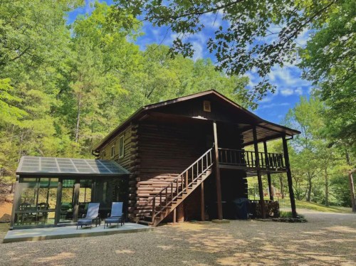 A rustic log cabin surrounded by trees, featuring a glass sunroom and a staircase leading to the upper level.
