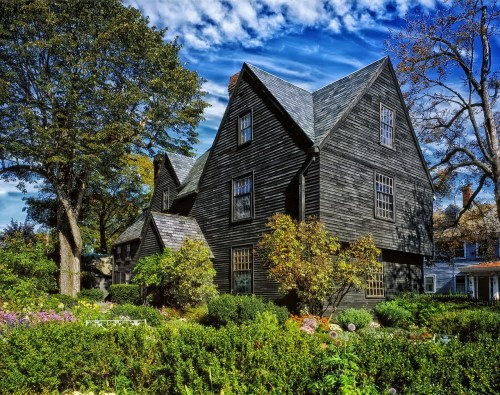 A large, dark wooden house with a unique roof design, surrounded by lush greenery and colorful flowers under a blue sky.