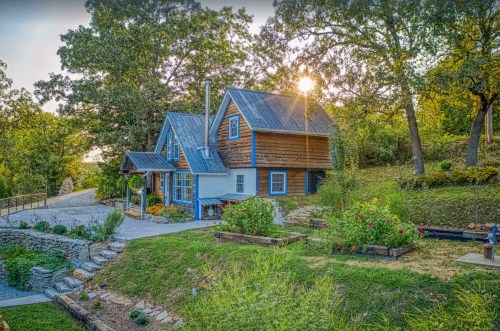 A charming wooden house with blue accents, surrounded by lush greenery and colorful flowers, under a sunset sky.