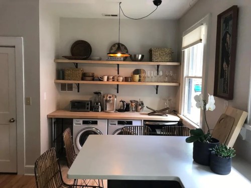 A modern kitchen with open shelves, a dining table, and laundry appliances, featuring plants and decorative items.