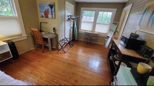 A cozy room with wooden floors, a desk, chair, and various furniture, featuring large windows and light-colored walls.
