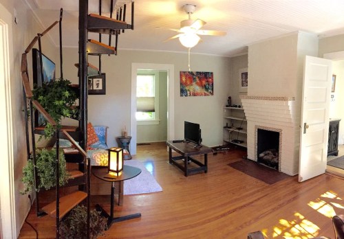 Cozy living room with a spiral staircase, wooden floors, a TV, and a fireplace, featuring plants and colorful artwork.
