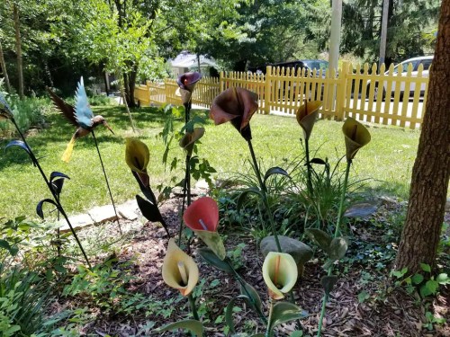Colorful metal flowers in a garden with a wooden fence and trees in the background on a sunny day.
