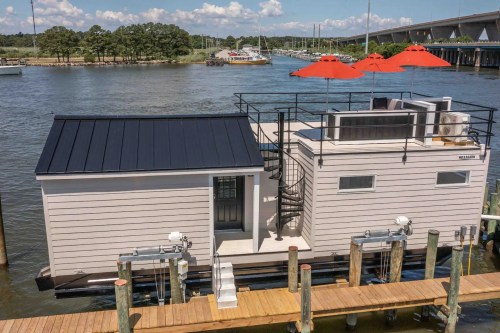 A modern houseboat with a black roof, spiral staircase, and red umbrellas, docked by a river with a bridge in the background.