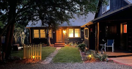 A cozy black house at dusk, warmly lit with a path leading to the front door, surrounded by greenery and outdoor seating.