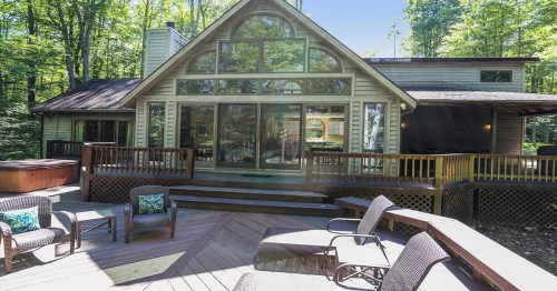 A modern cabin with large windows, surrounded by trees, featuring a deck and hot tub.