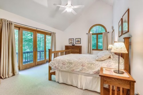 Bright bedroom with a wooden bed, nightstands, large windows, and a ceiling fan, surrounded by greenery.