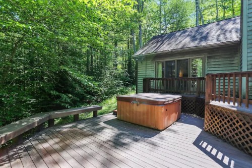 A wooden deck with a hot tub surrounded by lush green trees and a cozy cabin in the background.