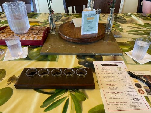 A table set for a tasting, featuring a flight of small glasses, water, a menu, and a platter of snacks.