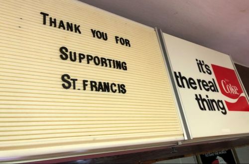Sign displaying "Thank you for Supporting St. Francis" next to a Coca-Cola advertisement saying "it's the real thing."