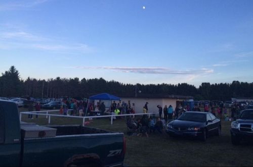 A crowd gathers near a building at dusk, with cars parked in a grassy area and a clear sky above.