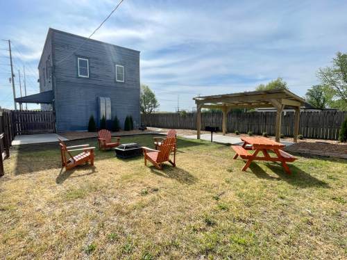 A backyard with a fire pit, picnic tables, and seating, surrounded by a wooden fence and a two-story building.