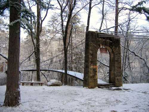 A snowy landscape featuring a stone archway and a suspension bridge surrounded by trees.