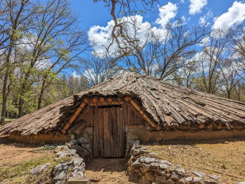 A rustic, thatched-roof hut surrounded by trees under a blue sky with scattered clouds.