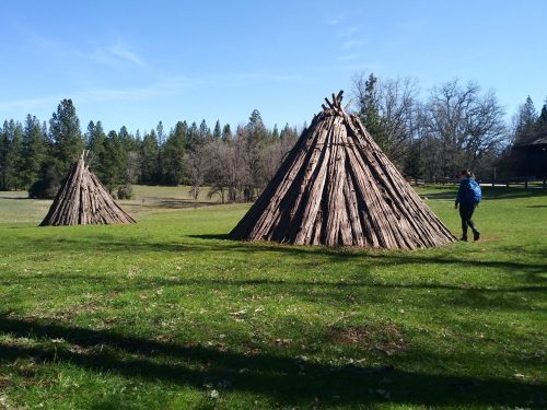 Two large teepee structures made of logs stand on a grassy field, with a person walking nearby under a clear blue sky.