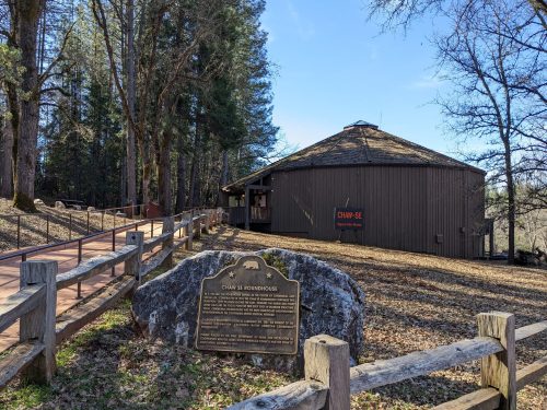 A round wooden building surrounded by trees, with a historical plaque in front and a clear blue sky above.