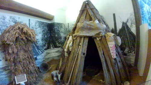 A traditional wooden structure resembling a teepee, surrounded by a mural of forested landscape.