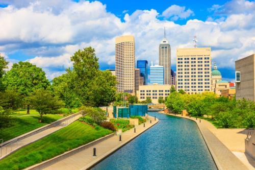 A scenic view of a city skyline with modern buildings beside a calm waterway and lush greenery under a blue sky.