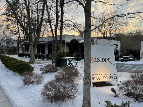 A snowy scene featuring the exterior of Indi-Q Indian Bistro & BBQ, with a sign and trees in the foreground.