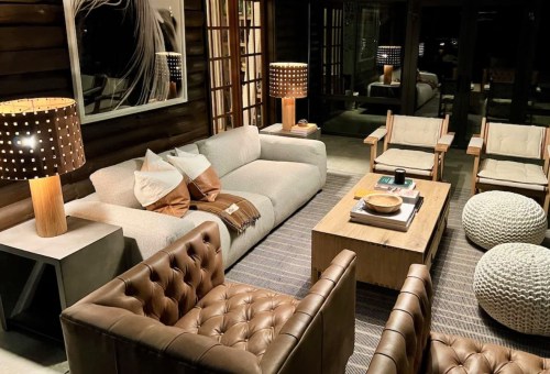 Cozy living room with a gray sofa, wooden coffee table, armchairs, and stylish lamps, featuring warm lighting and rustic decor.