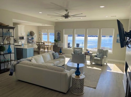 Bright, modern living room with white furniture, blue accents, and large windows overlooking a lake.