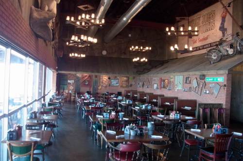 Interior of a rustic restaurant with wooden tables, chairs, and vintage decor, featuring chandeliers and large windows.