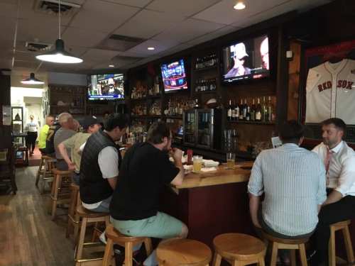 A cozy bar scene with patrons sitting at the counter, watching TVs, and enjoying drinks in a warm, inviting atmosphere.
