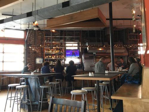 A cozy restaurant interior with patrons at the bar, wooden tables, and a large window letting in natural light.