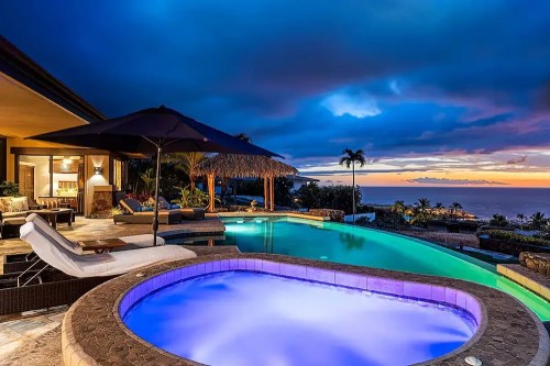 A luxurious villa with a pool and hot tub, overlooking a sunset-lit ocean and palm trees under a dramatic sky.