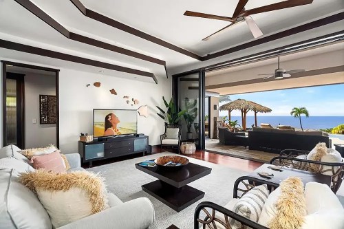 A modern living room with a TV, plush sofas, and ocean views through open sliding doors. Tropical decor and natural light.