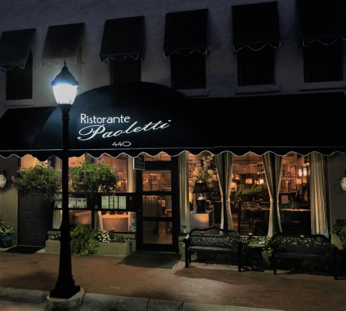 A cozy restaurant entrance at night, featuring an awning, outdoor seating, and warm lighting.